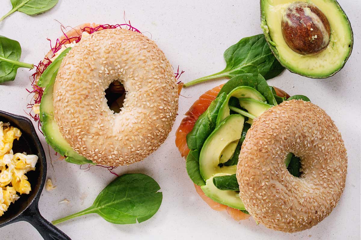 Sliced avocado neatly placed on a toasted bagel with toppings of microgreens and tomatoes.